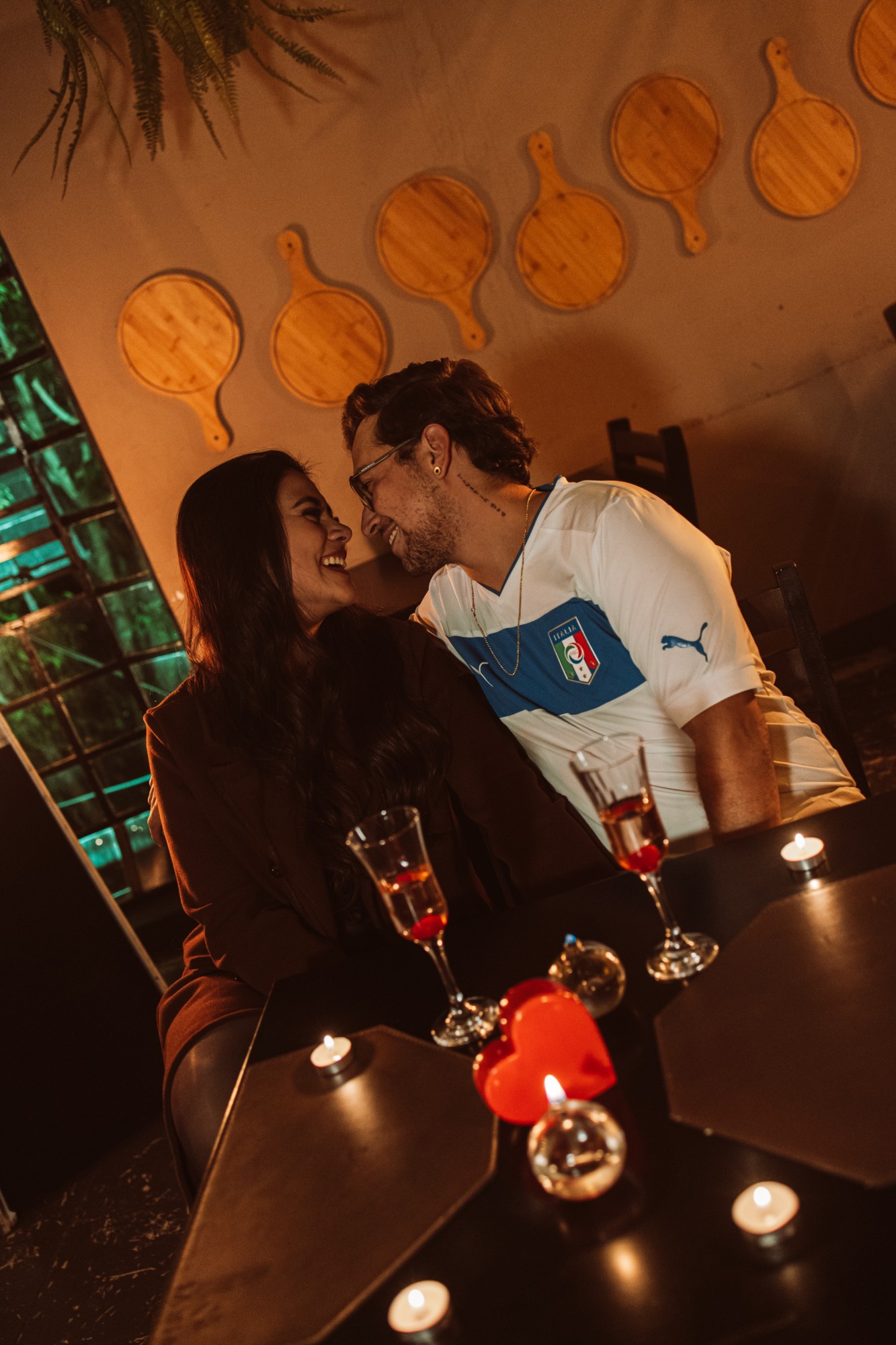 Casal rindo à mesa, com taças e velas, sob as tábuas de madeira da parede