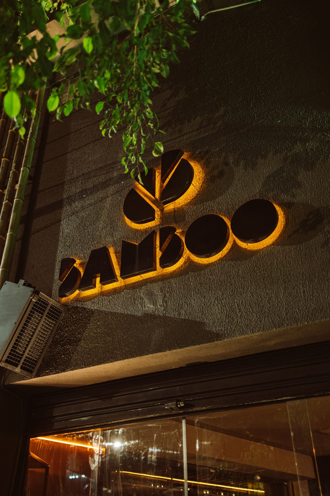 Letreiro iluminado do Bamboo na fachada, à noite, entre folhagens