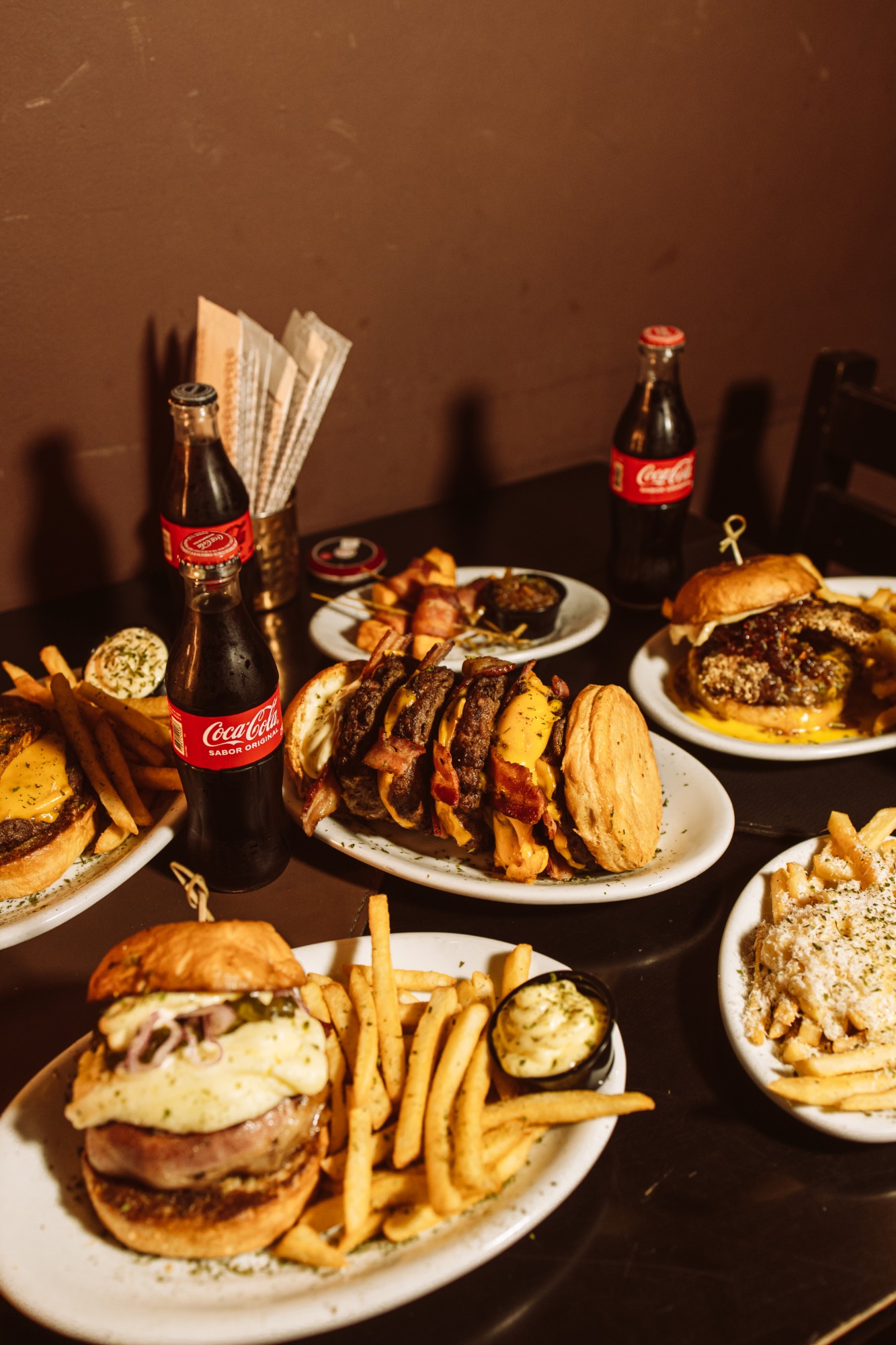 Mesa servida com vários hambúrgueres, porções de batata e refrigerantes